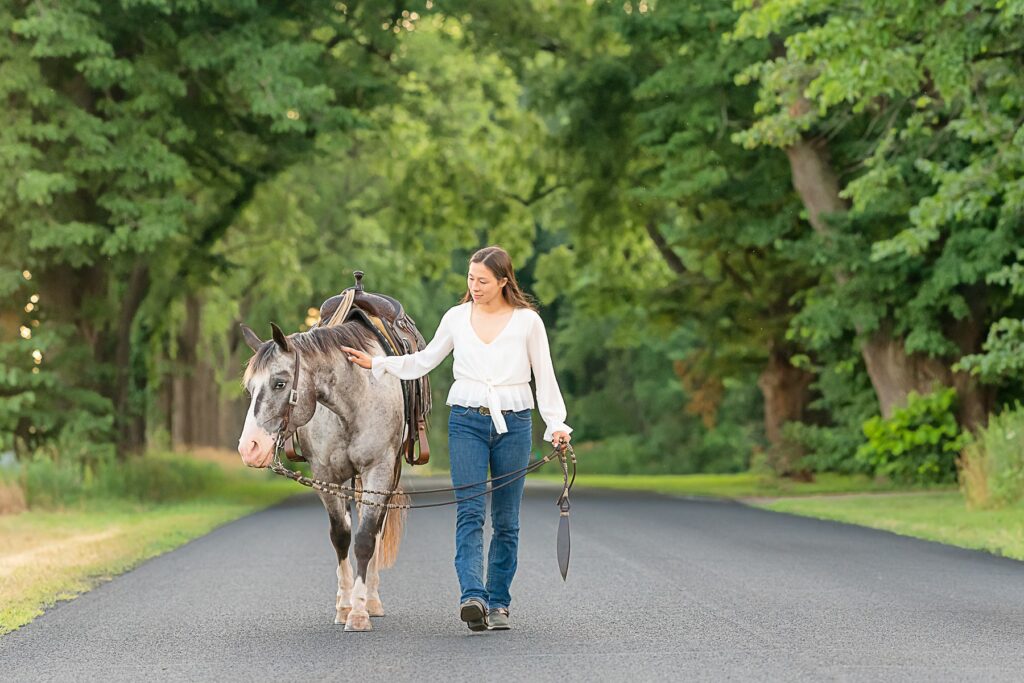 A teenage rider pets her Pony of the Americas on the shoulder while walking down a long, wooded driveway. Quiet roads make scenic options for equestrian portraits