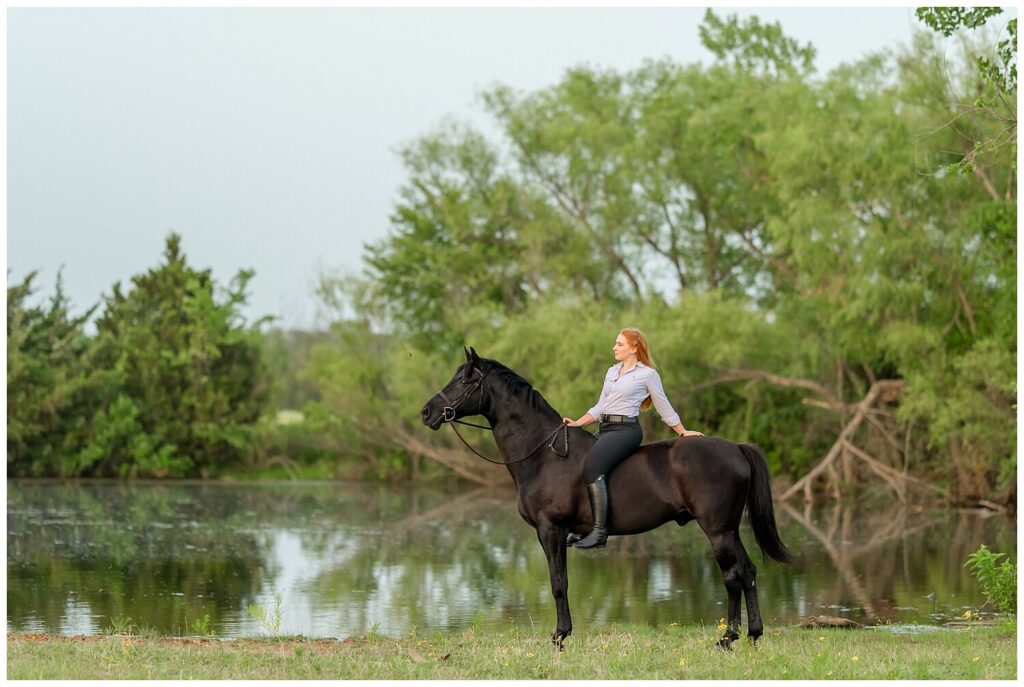 A redheaded rider sits bareback on her black hunter gelding as they stand in front of a pond. Ponds and other water elements add romanticism to your equestrian photo shoot