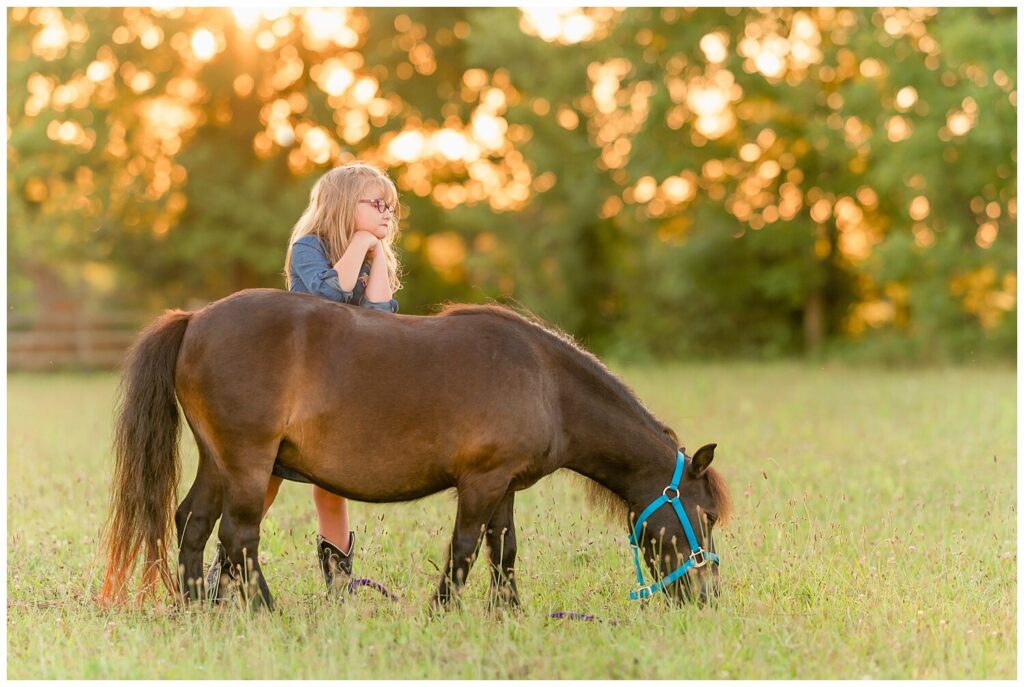 A young girl leans on the back of a bay miniature horse as he grazes in a pasture. Your horse's pasture is a good location option for your equestrian portrait session 