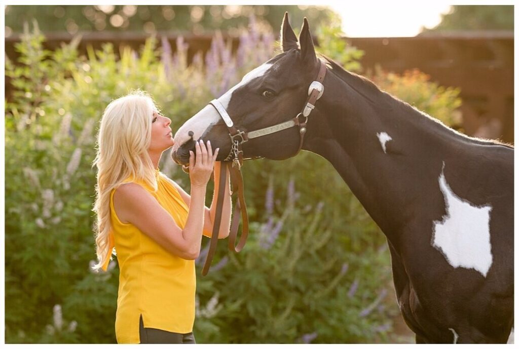 A woman kisses her Paint Horse in front of flowering bushes at her barn. Choosing scenic locations close to your horse's home can help him stay calm