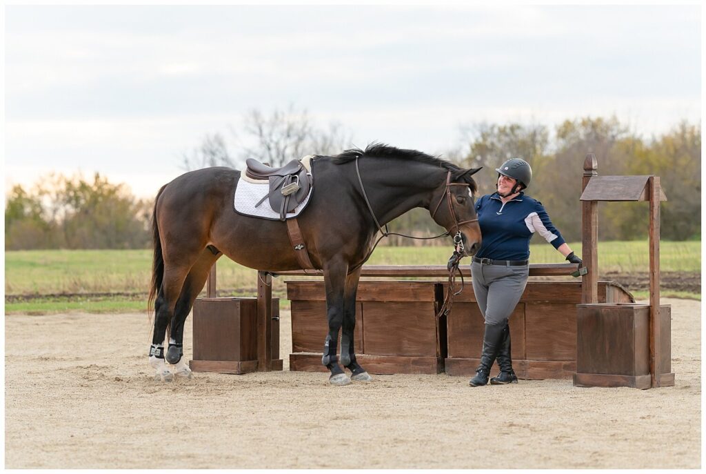An equestrian stands with her hunter gelding in full tack and attire in front of a brown wishing well roll top jump. Arenas are good locations for horse and rider portrait sessions.