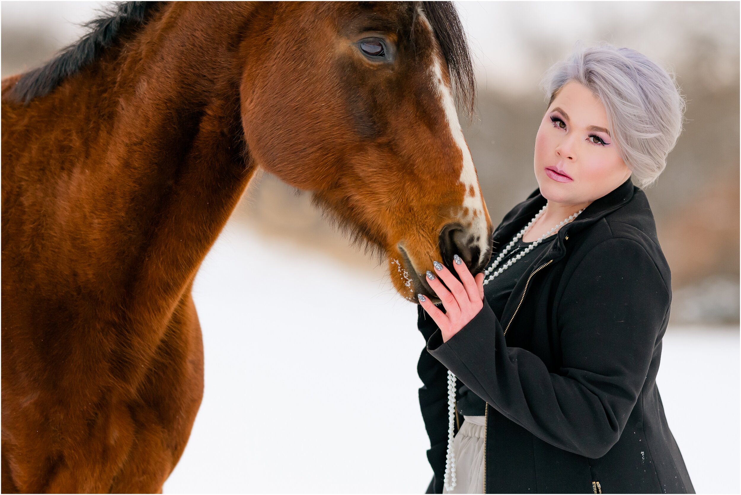  Girl with grey hair stands in the snow in a black winter wool coat alongside  her senior quarter horse mare.  Grey skirt outfit for equestrian portrait session 