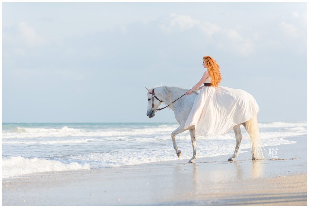 A redheaded woman in a long white dress walks a grey Andalusian mare into the ocean waves during an equestrian portrait session 