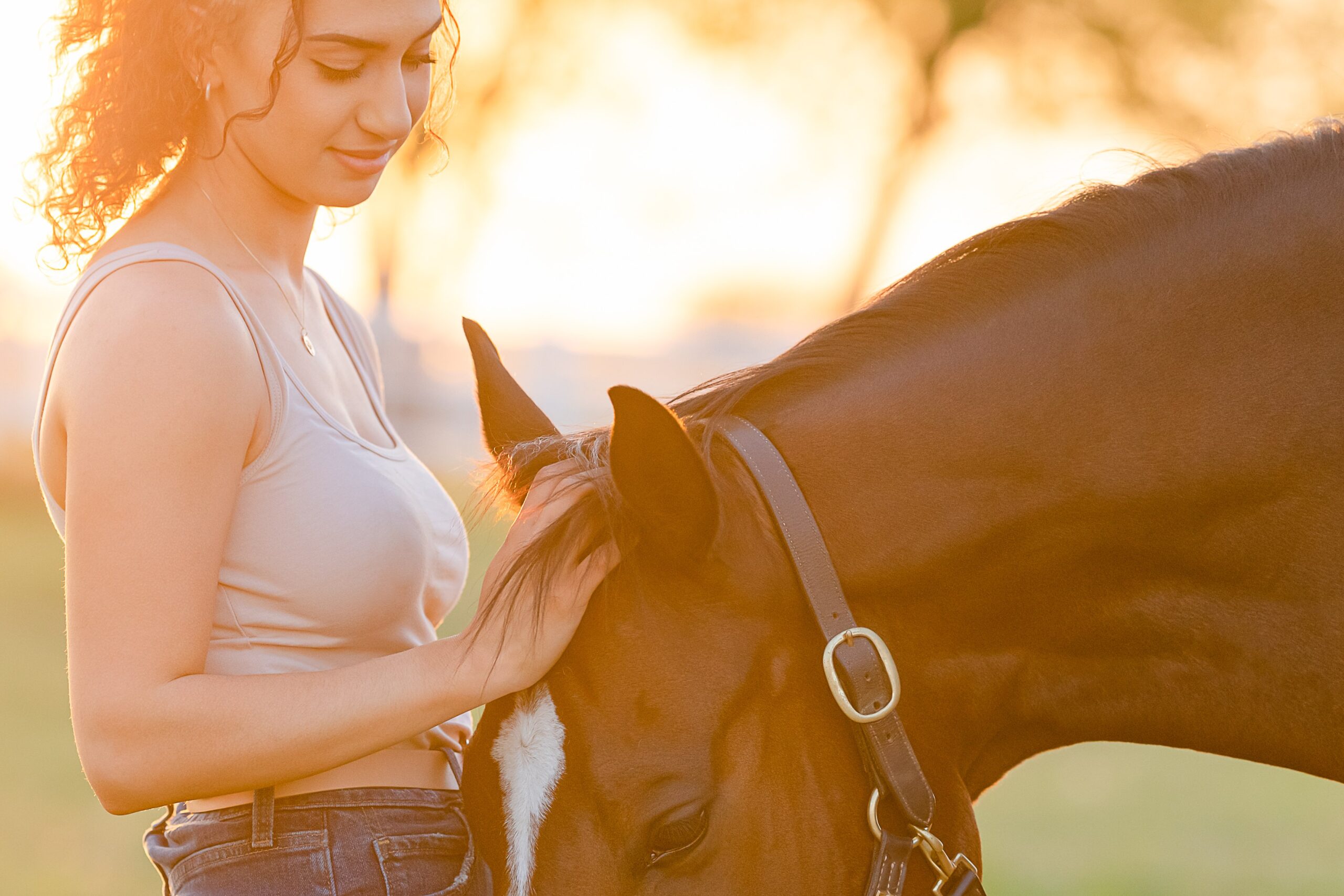 A 20-something woman with curly hair looks down at her horse's lowered head and pets his forelock