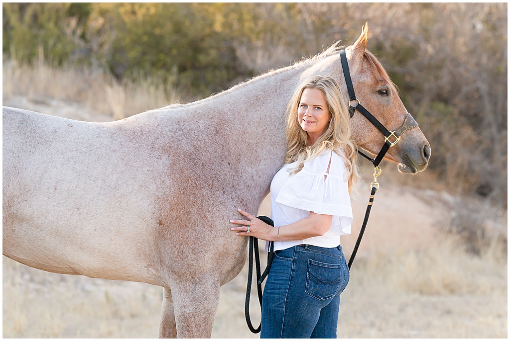 a blonde woman in blue jeans and a white shirt poses with her red roan quarter horse mare in Big Spring Texas