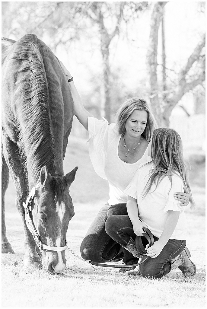 A black and white image of a mother and daughter crouching near their grazing horse. the mother is gazing at her daughter