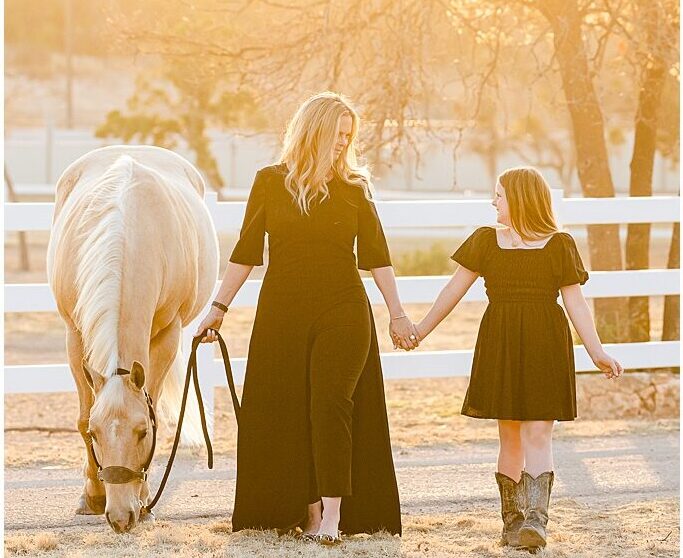 A mother and daughter hold hands at sunset while leading their palomino quarter horse