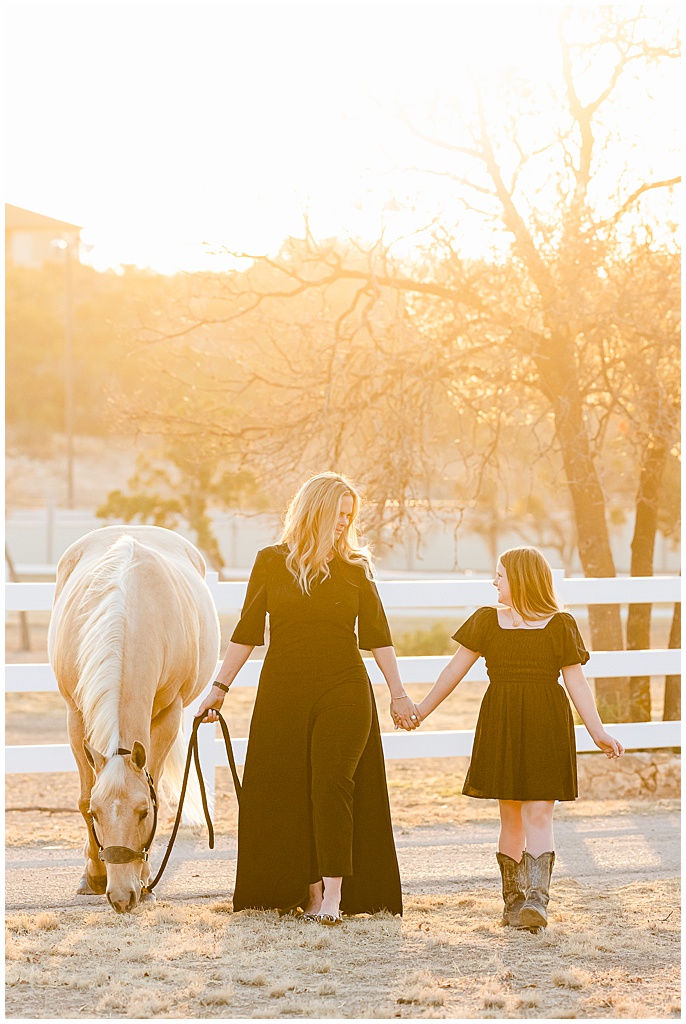 A mother and daughter hold hands at sunset while leading their palomino quarter horse